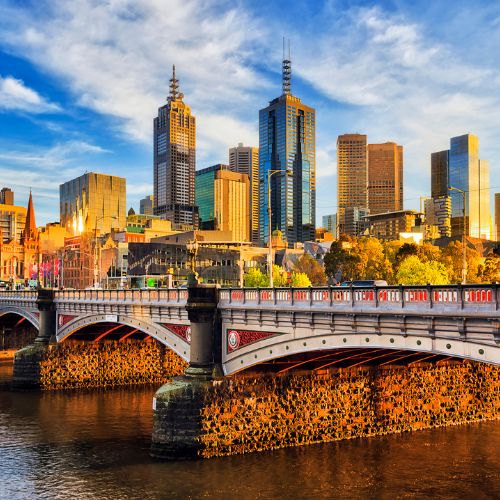A vibrant cityscape of Melbourne at sunset, featuring skyscrapers, a historic church, and a detailed arched bridge reflecting in the Yarra River.