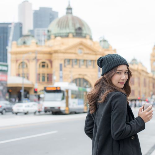Woman in a black coat and beanie stands on a busy city street, holding a phone. Iconic building and tram in the background. Daytime, lively urban vibe.
