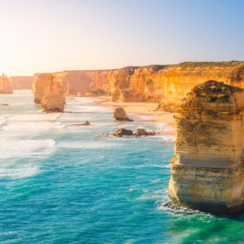 Sunlit coastal scene of the Twelve Apostles in Australia. Eroded limestone stacks rise from blue ocean waves with a backdrop of rugged cliffs.