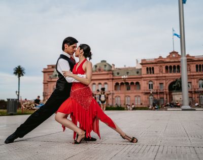 A couple dances passionately, performing a tango pose in front of a historic pink building. The woman wears a red dress, and the man is in black attire.
