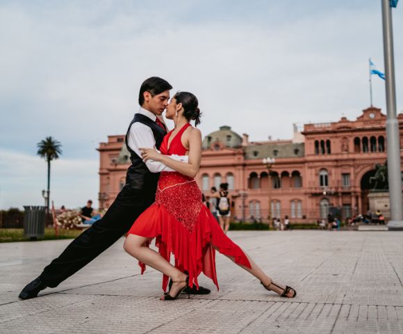 A couple dances passionately, performing a tango pose in front of a historic pink building. The woman wears a red dress, and the man is in black attire.