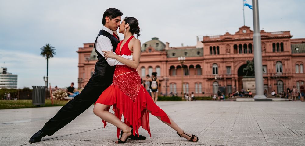 A couple dances passionately, performing a tango pose in front of a historic pink building. The woman wears a red dress, and the man is in black attire.
