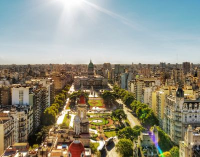Aerial view of a bustling cityscape featuring a historic building with a green dome surrounded by trees and parks, under a clear, sunny sky.