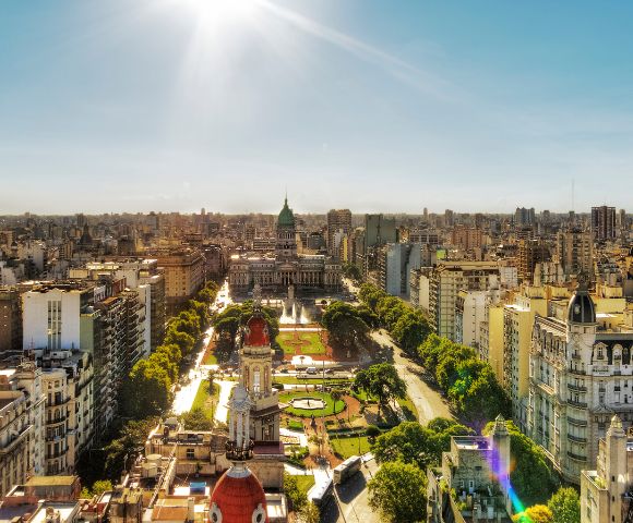 Aerial view of a bustling cityscape featuring a historic building with a green dome surrounded by trees and parks, under a clear, sunny sky.