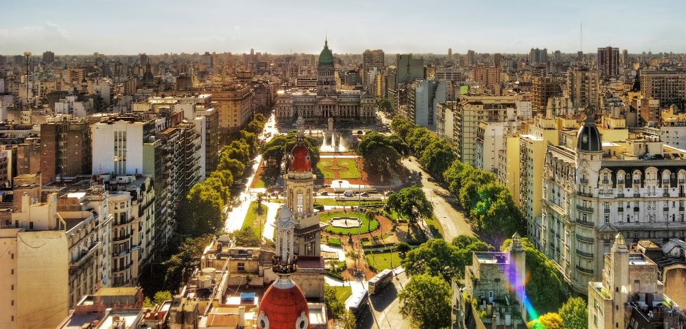 Aerial view of a bustling cityscape featuring a historic building with a green dome surrounded by trees and parks, under a clear, sunny sky.