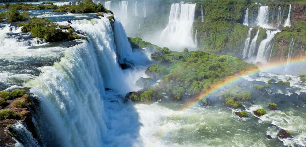 Majestic waterfalls cascade down lush green cliffs, with mist rising into the air. A vibrant rainbow arcs over the scene, creating a serene, awe-inspiring view.