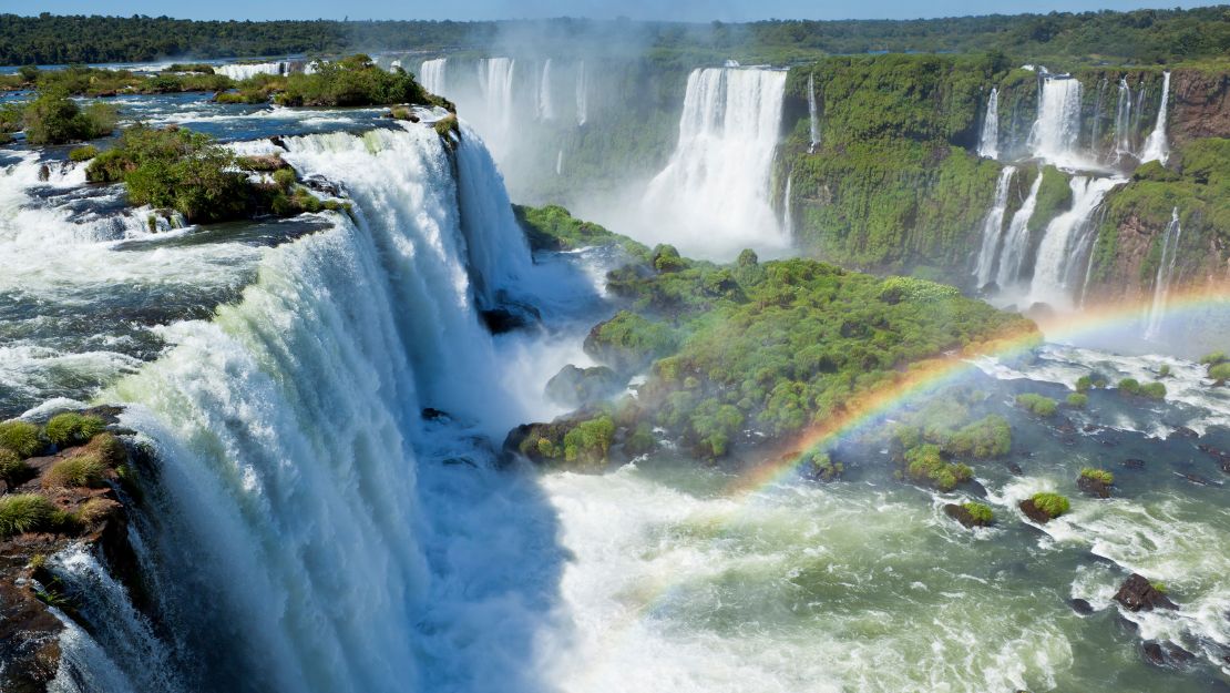 Majestic waterfalls cascade down lush green cliffs, with mist rising into the air. A vibrant rainbow arcs over the scene, creating a serene, awe-inspiring view.