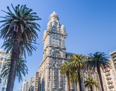 A tall, ornate building with a unique design stands against a clear blue sky, surrounded by towering palm trees, creating a vibrant and majestic urban scene.