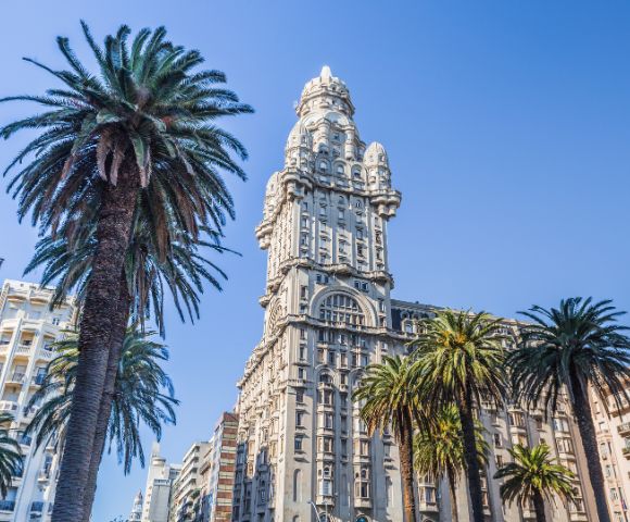 A tall, ornate building with a unique design stands against a clear blue sky, surrounded by towering palm trees, creating a vibrant and majestic urban scene.