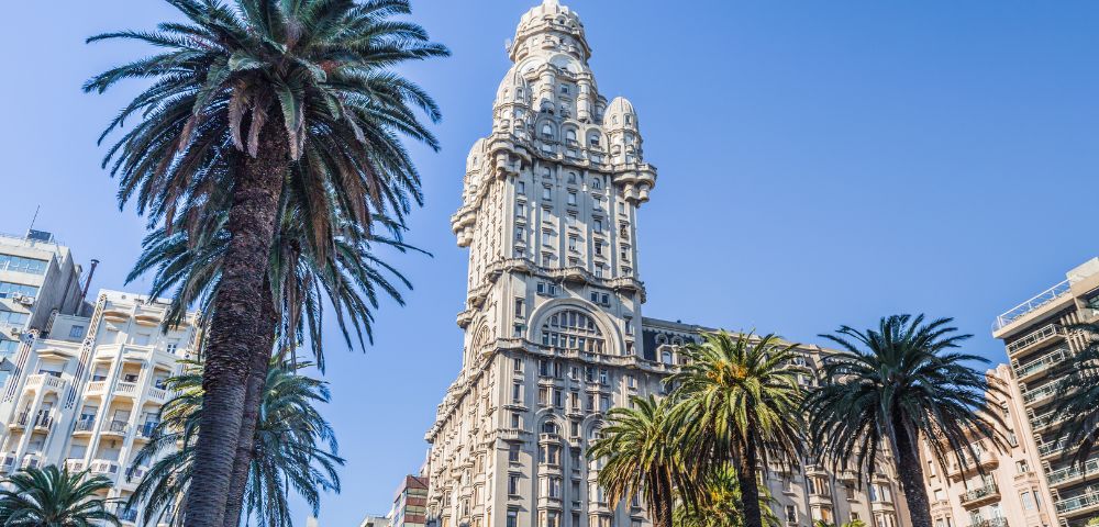 A tall, ornate building with a unique design stands against a clear blue sky, surrounded by towering palm trees, creating a vibrant and majestic urban scene.