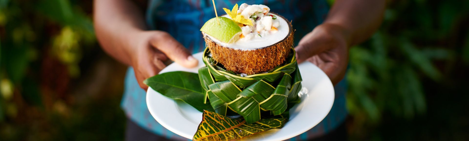 Person holding a white plate with a coconut shell filled with a dish, garnished with lime and leaves, surrounded by lush greenery, conveying a tropical vibe.