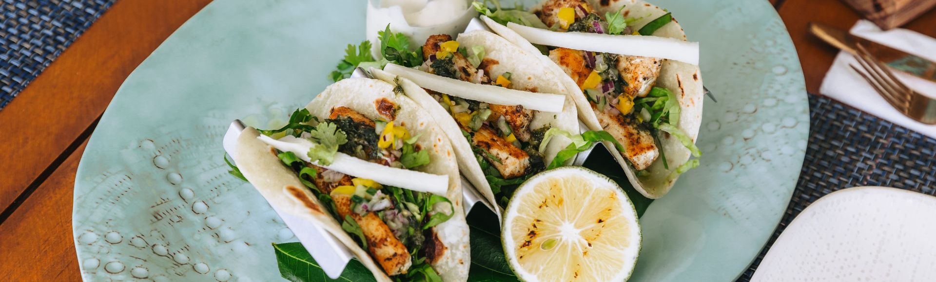 Three tacos on a light blue plate, filled with grilled chicken, diced vegetables, and fresh cilantro, accompanied by a halved lemon. The presentation is fresh and colorful.