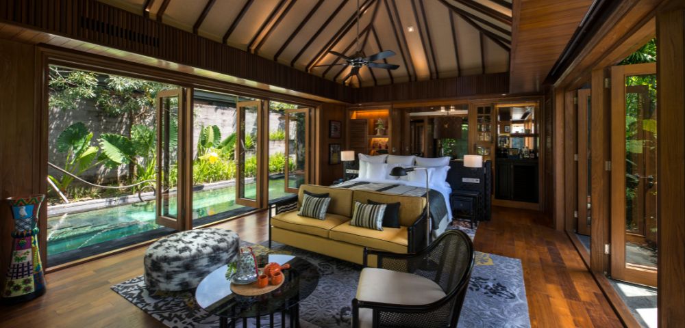 Luxurious tropical bedroom with wood accents, high ceilings, and large windows overlooking lush greenery. Cozy seating, elegant decor, serene ambiance.