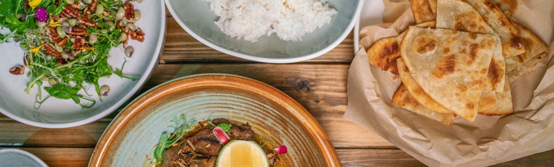 Aerial view of a wooden table with a vibrant salad, fluffy white rice, crispy flatbread, and a hearty meat dish garnished with a lemon slice, conveying a warm and inviting meal.