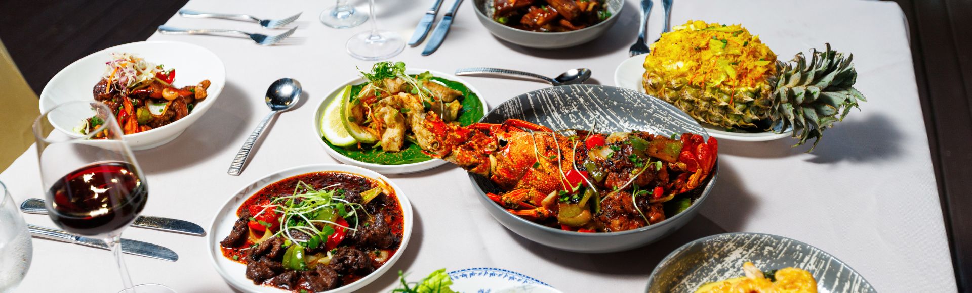 An elegant dining table with various dishes, including a whole fish, stir-fried vegetables, and pineapple fried rice. A glass of red wine is visible, creating a vibrant, appetizing scene.