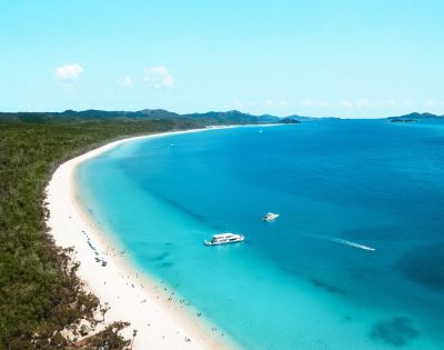 Aerial view of a serene beach with white sand and turquoise water. Two boats float offshore. Lush green hills line the horizon under a clear blue sky.