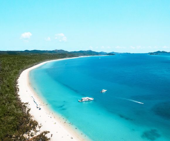 Aerial view of a serene beach with white sand and turquoise water. Two boats float offshore. Lush green hills line the horizon under a clear blue sky.