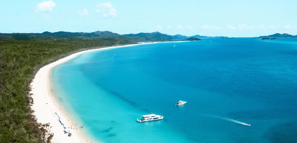 Aerial view of a serene beach with white sand and turquoise water. Two boats float offshore. Lush green hills line the horizon under a clear blue sky.