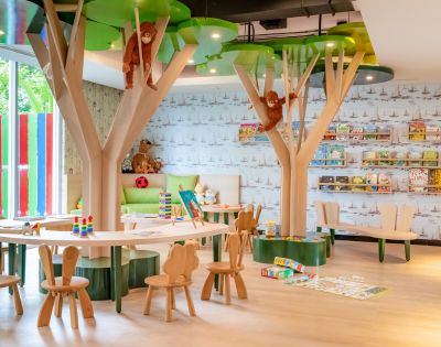 Children’s playroom with tree-themed decor, small wooden chairs, and tables. Plush monkeys hang from tree structures. Toys and books line the walls, creating a vibrant and inviting atmosphere.