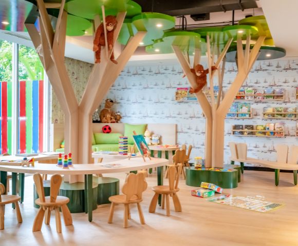 Children’s playroom with tree-themed decor, small wooden chairs, and tables. Plush monkeys hang from tree structures. Toys and books line the walls, creating a vibrant and inviting atmosphere.