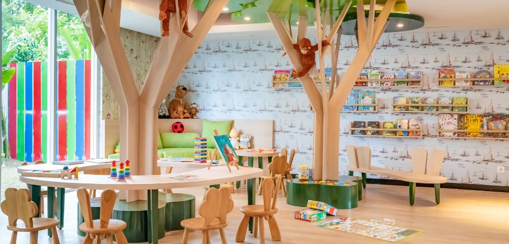 Children’s playroom with tree-themed decor, small wooden chairs, and tables. Plush monkeys hang from tree structures. Toys and books line the walls, creating a vibrant and inviting atmosphere.