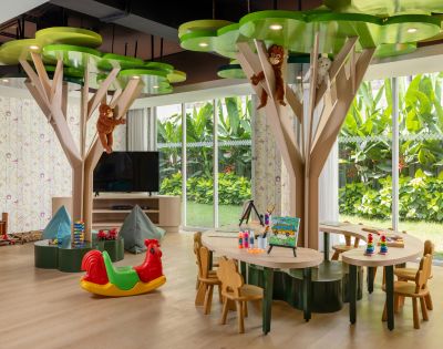 Bright children's playroom with tree-shaped tables, monkey decorations, and toys. Large windows show lush greenery outside, creating a joyful atmosphere.