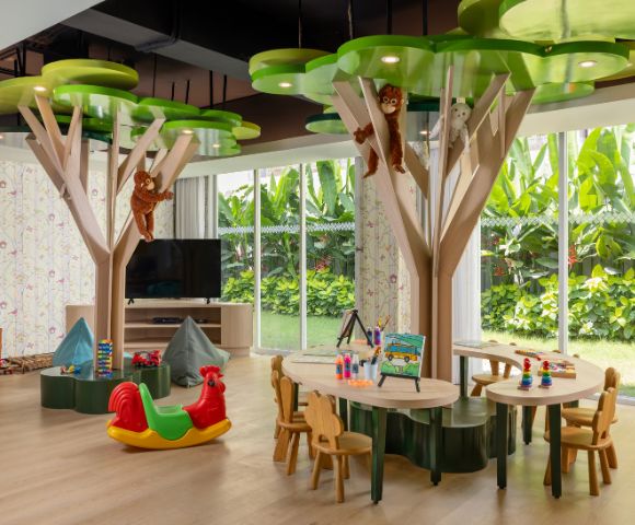 Bright children's playroom with tree-shaped tables, monkey decorations, and toys. Large windows show lush greenery outside, creating a joyful atmosphere.