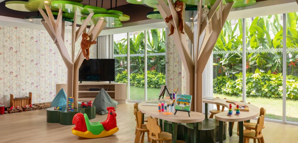 Bright children's playroom with tree-shaped tables, monkey decorations, and toys. Large windows show lush greenery outside, creating a joyful atmosphere.