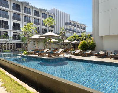Luxurious hotel pool area with blue tiles, surrounded by sun loungers and umbrellas. Modern multi-story building in the background under a clear blue sky.