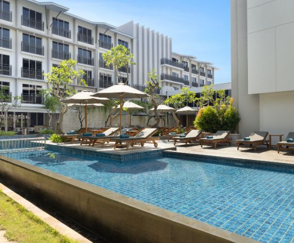 Luxurious hotel pool area with blue tiles, surrounded by sun loungers and umbrellas. Modern multi-story building in the background under a clear blue sky.