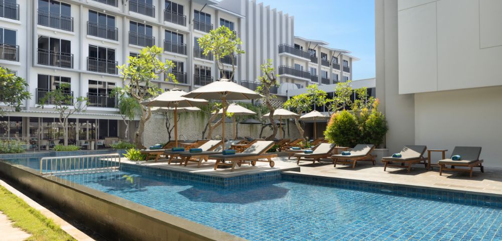 Luxurious hotel pool area with blue tiles, surrounded by sun loungers and umbrellas. Modern multi-story building in the background under a clear blue sky.