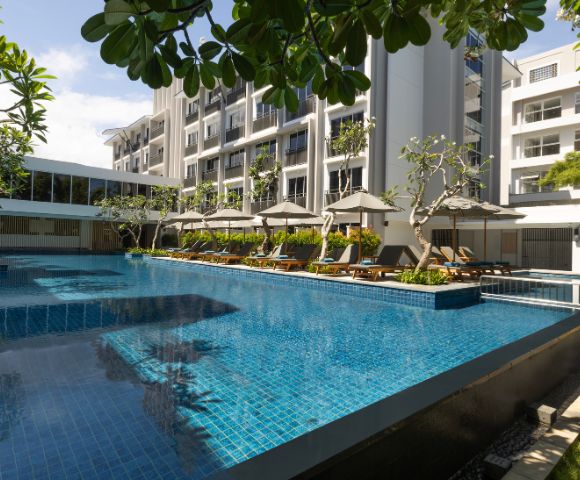 Luxurious hotel pool with clear blue water, surrounded by modern white buildings. Sun loungers with umbrellas line the poolside, creating a serene setting.