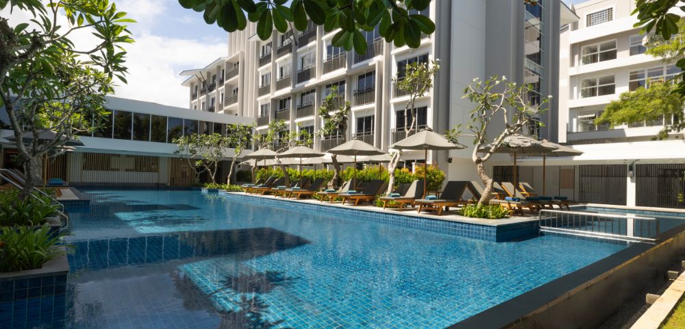 Luxurious hotel pool with clear blue water, surrounded by modern white buildings. Sun loungers with umbrellas line the poolside, creating a serene setting.