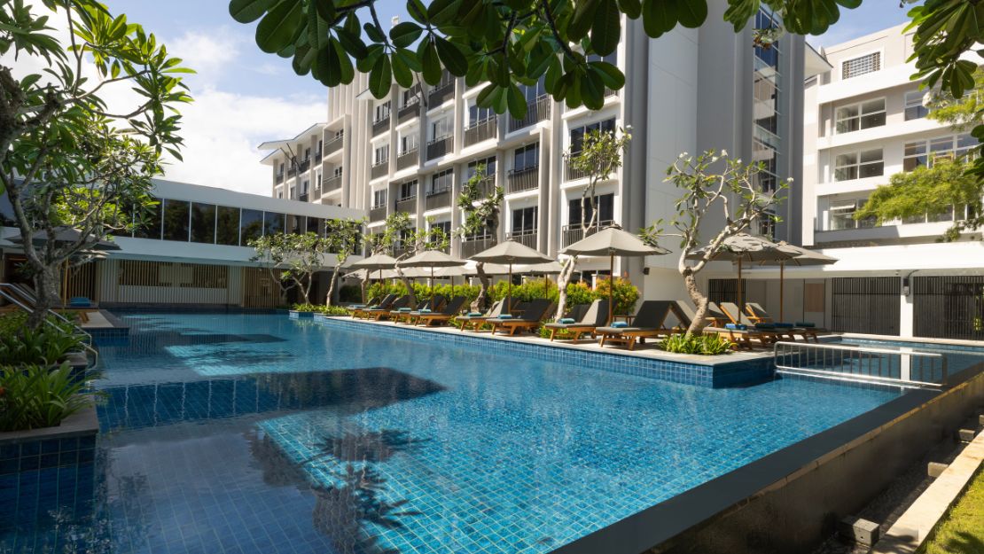 Luxurious hotel pool with clear blue water, surrounded by modern white buildings. Sun loungers with umbrellas line the poolside, creating a serene setting.