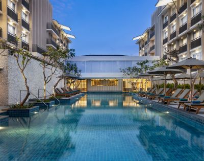 Luxurious hotel pool surrounded by lounge chairs and umbrellas at dusk, flanked by modern buildings with balconies. The serene evening ambiance is accentuated by soft lighting.
