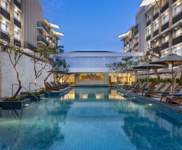 Luxurious hotel pool surrounded by lounge chairs and umbrellas at dusk, flanked by modern buildings with balconies. The serene evening ambiance is accentuated by soft lighting.