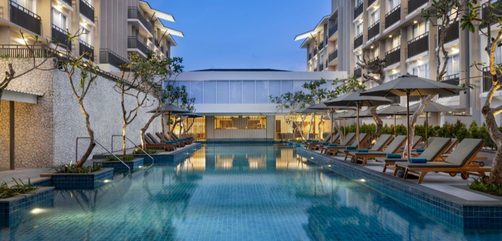 Luxurious hotel pool surrounded by lounge chairs and umbrellas at dusk, flanked by modern buildings with balconies. The serene evening ambiance is accentuated by soft lighting.