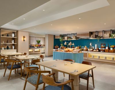 Modern restaurant interior with light wood tables, blue cushioned chairs, and a buffet counter showcasing varied dishes. Ambient lighting creates a cozy atmosphere. Blue accent wall and decorative shelves enhance the elegant design.