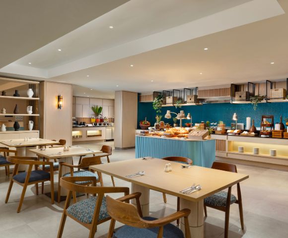 Modern restaurant interior with light wood tables, blue cushioned chairs, and a buffet counter showcasing varied dishes. Ambient lighting creates a cozy atmosphere. Blue accent wall and decorative shelves enhance the elegant design.