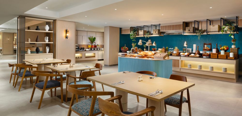 Modern restaurant interior with light wood tables, blue cushioned chairs, and a buffet counter showcasing varied dishes. Ambient lighting creates a cozy atmosphere. Blue accent wall and decorative shelves enhance the elegant design.