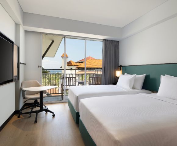 Modern hotel room with two neatly made beds, a flat-screen TV, desk with chair, and a balcony with a view of terracotta-roofed buildings. Bright and inviting.