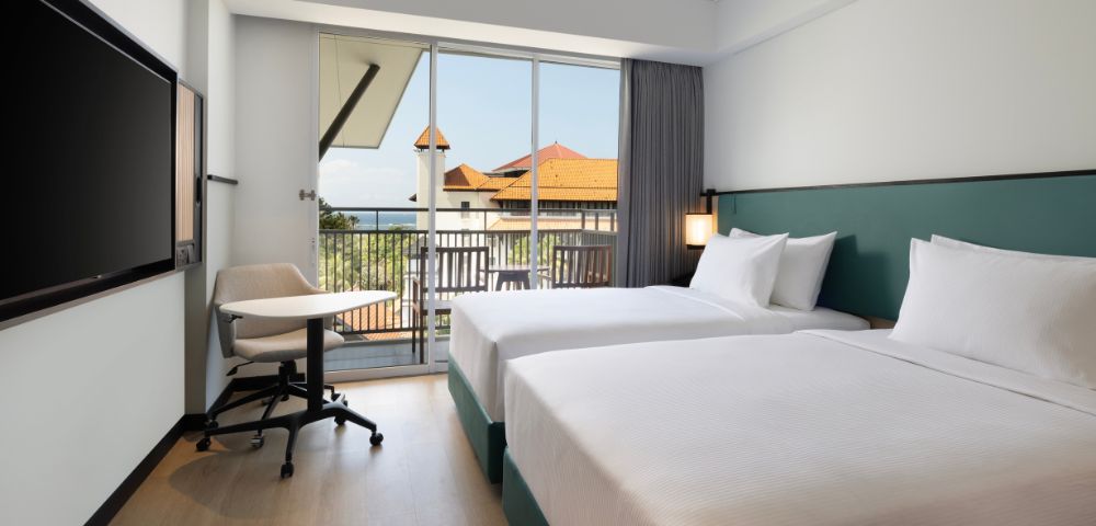Modern hotel room with two neatly made beds, a flat-screen TV, desk with chair, and a balcony with a view of terracotta-roofed buildings. Bright and inviting.