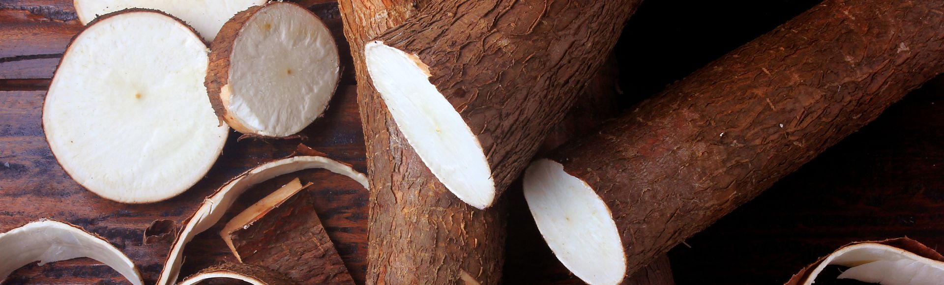 Alt text: Fresh cassava roots with rough brown skin, some sliced to reveal the white flesh inside, arranged on a wooden surface with peeled strips nearby.