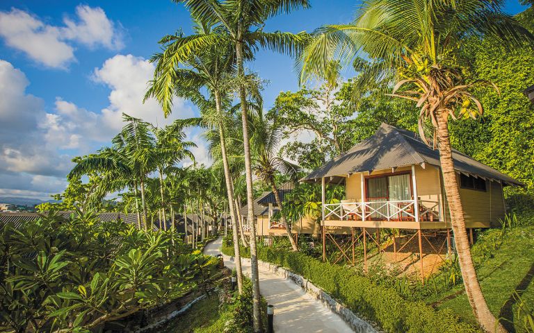 Tropical resort scene with thatched-roof bungalows on stilts, surrounded by lush greenery and palm trees under a partly cloudy blue sky, conveying tranquility.