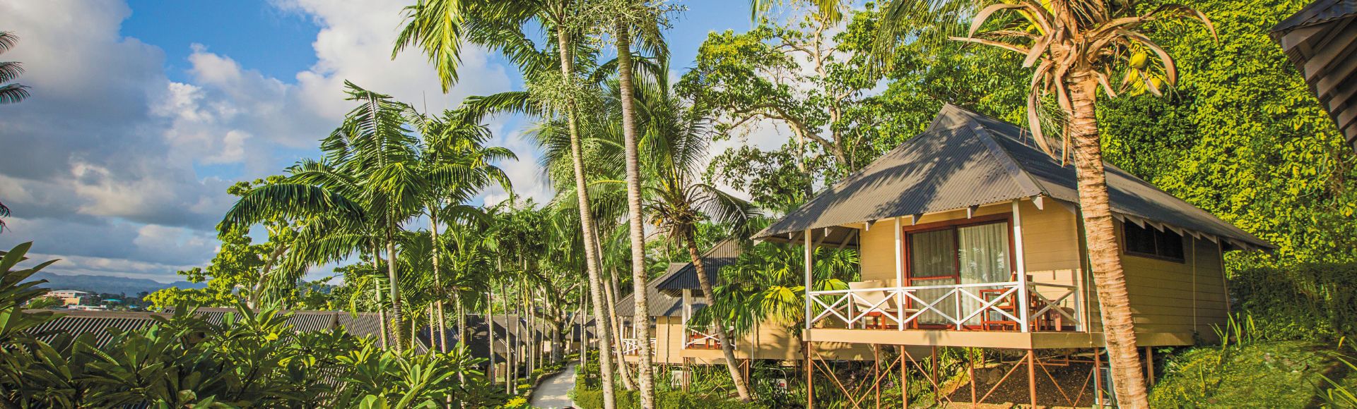 Tropical resort scene with thatched-roof bungalows on stilts, surrounded by lush greenery and palm trees under a partly cloudy blue sky, conveying tranquility.