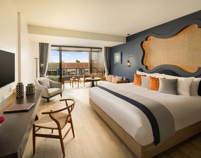 Modern hotel room with a large bed, vibrant cushions, dark blue accent wall, and a wall-mounted TV. Glass doors open to a balcony with seating, overlooking terracotta rooftops under a clear sky. Room exudes warmth and comfort.