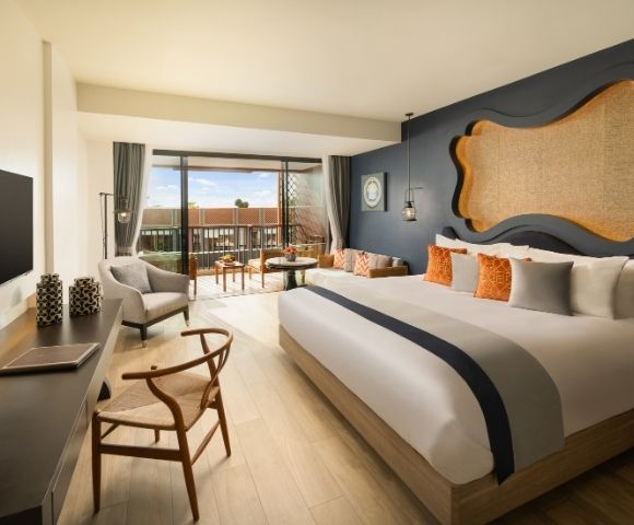 Modern hotel room with a large bed, vibrant cushions, dark blue accent wall, and a wall-mounted TV. Glass doors open to a balcony with seating, overlooking terracotta rooftops under a clear sky. Room exudes warmth and comfort.