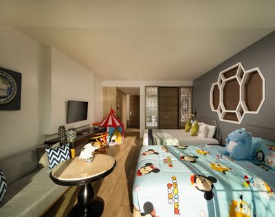 Bright hotel room with playful decor featuring cartoon-themed bedding, a blue plush toy, a red play tent, and modern furnishings on a wooden floor.