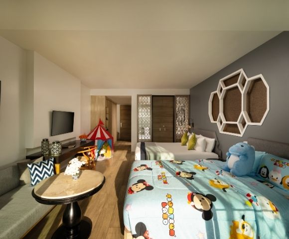 Bright hotel room with playful decor featuring cartoon-themed bedding, a blue plush toy, a red play tent, and modern furnishings on a wooden floor.