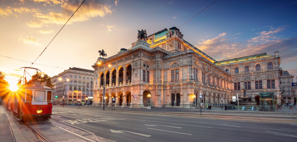 A historic opera house in Vienna is illuminated by the warm glow of sunset. A vintage tram passes in the foreground, evoking a sense of nostalgia and elegance.